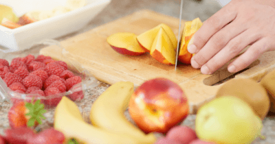 Man sieht ein Holzschneidebrett, Hände die einen Pfirsich schneiden, zwei Bananen, Pfirsich, Apfel, Kiwi, Erdbeeren und eine Schale Himbeeren.