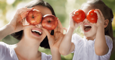 Man sieht eine Mama mit je einem Apfel vor den Augen und ein Kind mit zwei Tomaten sich vor die Augen halten. Beide tragen weisse T-Shirts.