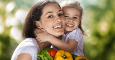 Man sieht eine Mama mit ihrer Tochter Wange an Wange. Beide lachen und tragen ein weisses T-Shirt. Im Hintergrund ist grün verschwommen etwas zu sehen. In der Hand der Mutter sind Salat, Tomate und zwei gelbe Peperoni zu sehen!