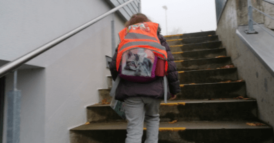 Man sieht ein Kind mit Rucksack und Weste die Treppe zum Schulhaus rauflaufen.