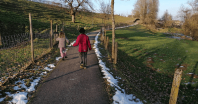 Man sieht zwei Kinder entlang eines Weges laufen. Links und rechts ist Wiese und ein Zaun.