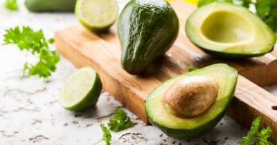 Man sieht Avocado, ganz und halbiert auf einem Holzbrett. Es sind auch grüne Limetten zu sehen. Kochen und Braten ohne Fett oder Öl