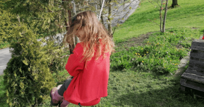Man sieht ein Kind mit einer roten Jacke auf einer Schaukel im Garten. Sie hat lange dunkelblonde Haare. Im Hintergrund ist der Garten zu sehen. 12 von 12 im April 2022.