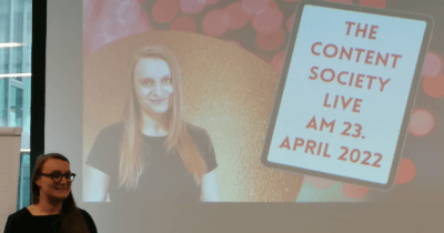 Man sieht Judith Peters vor einer Leinwand stehen. Da ist ein Bild von ihr zu sehen mit dem Text: The Content Society Live am 23. April 2022.