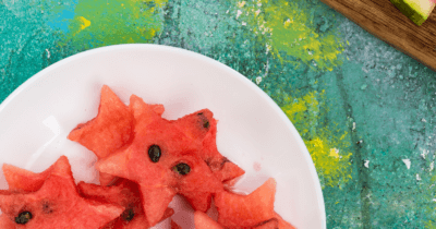 Man sieht Wassermelonen-Sterne auf einem weissen Teller. Dieser steht auf einem grün, gelb bunten Tisch. Es ist noch wenig von einem Holzschneidebrett und Wassermelonen-Schalen zu sehen.