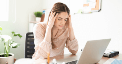 Man sieht eine gestresste Frau in ihrem Büro vor dem Computer sitzen. Sie hat den Kopf in die Hände gestützt.